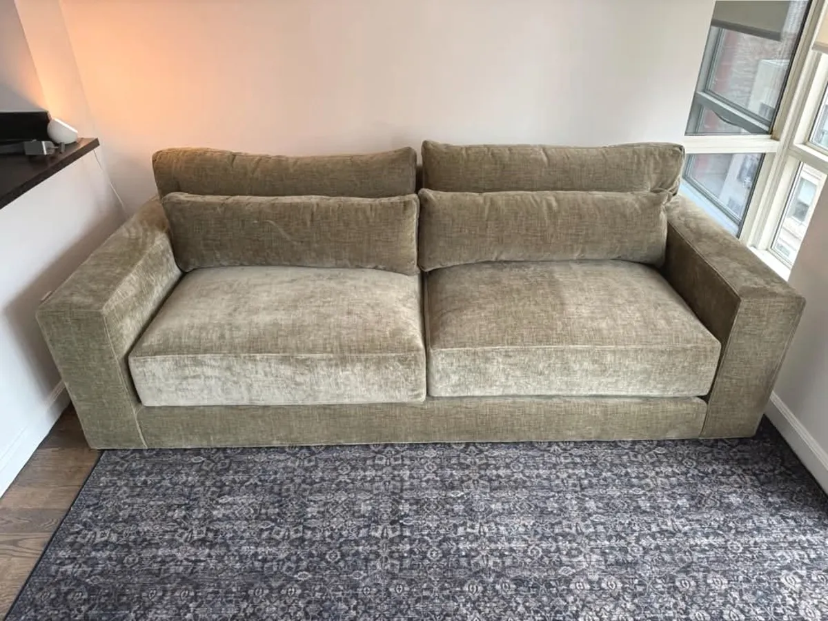 A sage velvet two-seat sofa on a grey patterned rug between two windows