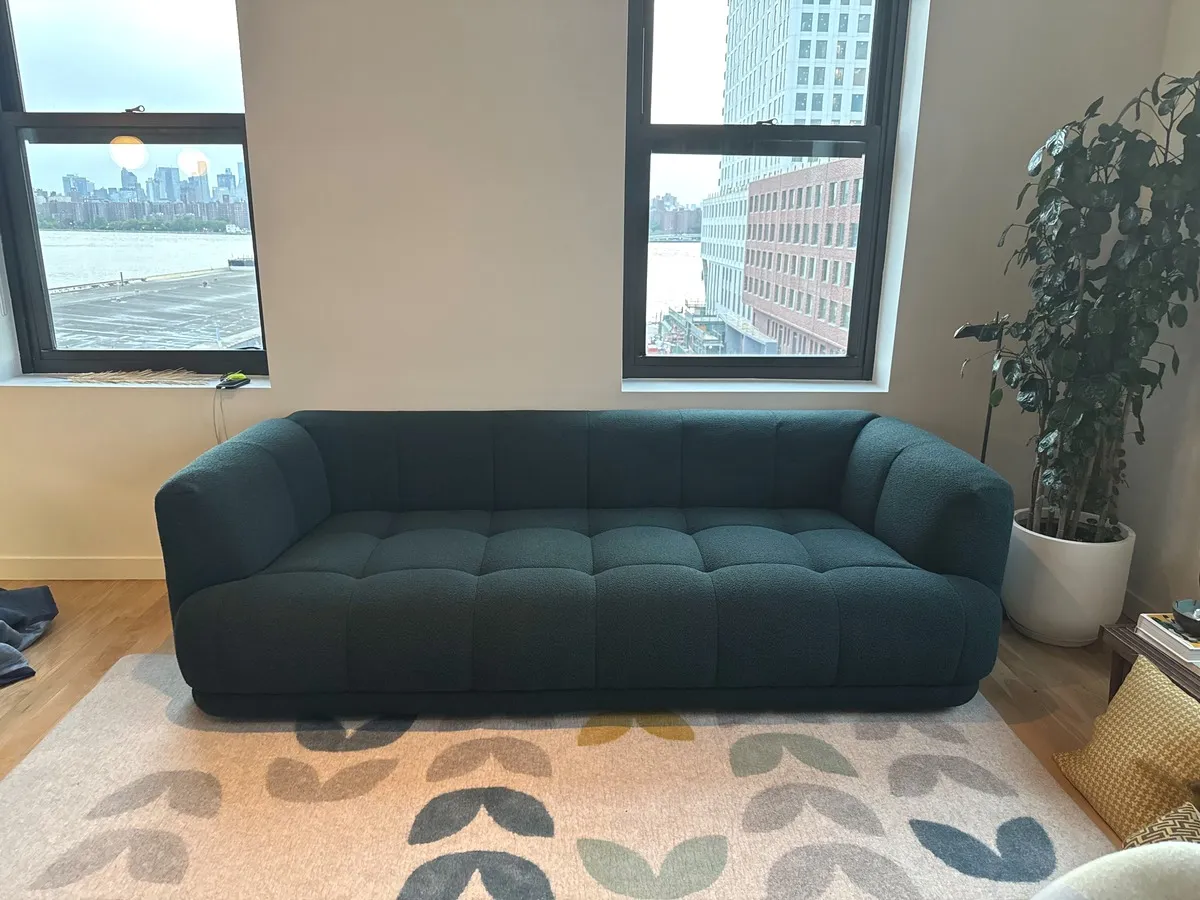 A low tufted teal sofa in a waterfront apartment, framed by two windows