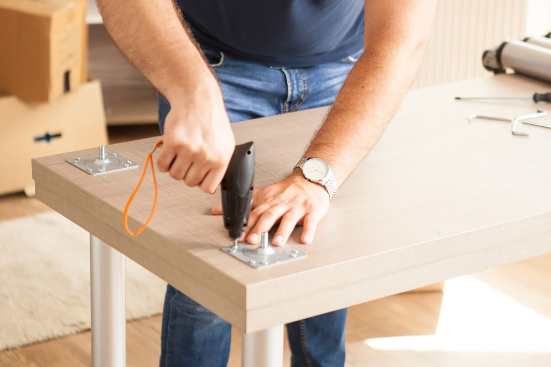 Person using a power drill to attach a metal bracket to a wooden table—furniture assembly in progress.