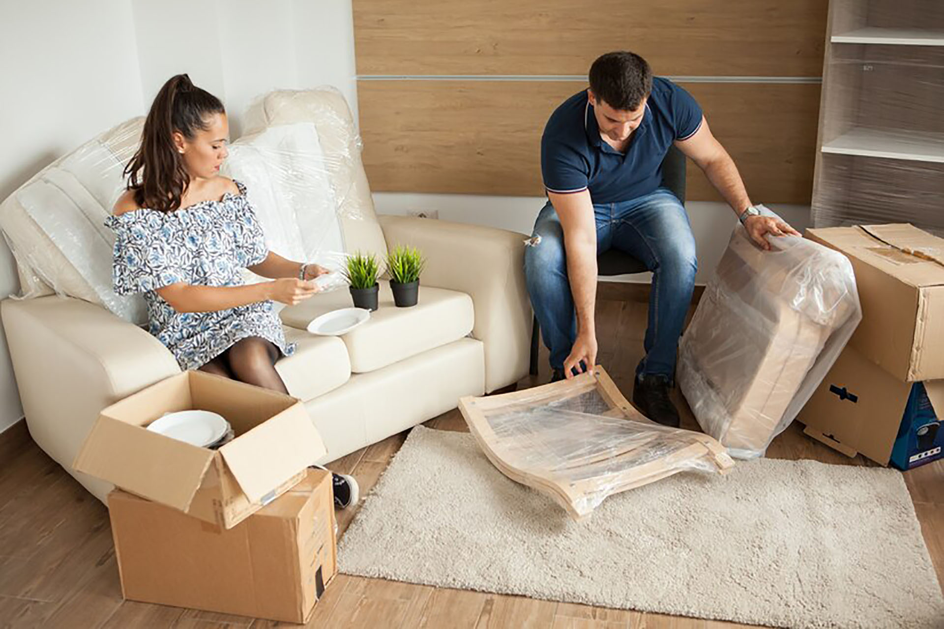 Two people unpacking in a cozy room with boxes, a white couch, and potted plants—settling into a new home.